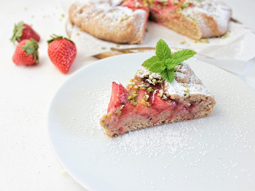 Erdbeerkuchenstück mit Minzgarnitur auf weißem Teller, frische Erdbeeren im Hintergrund, mit Puderzucker bestreut.