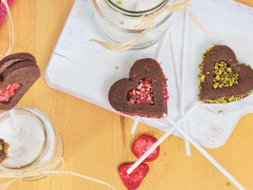 Herzförmige Schokoladen-Lollis mit buntem Dekor, auf Holzbrett angerichtet, daneben getrocknete Erdbeeren.