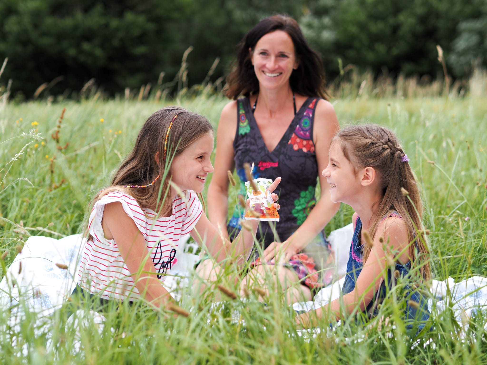 Mutter und Töchter genießen ein Picknick im Grünen. Lächelnde Gesichter in einer Sommerwiese.