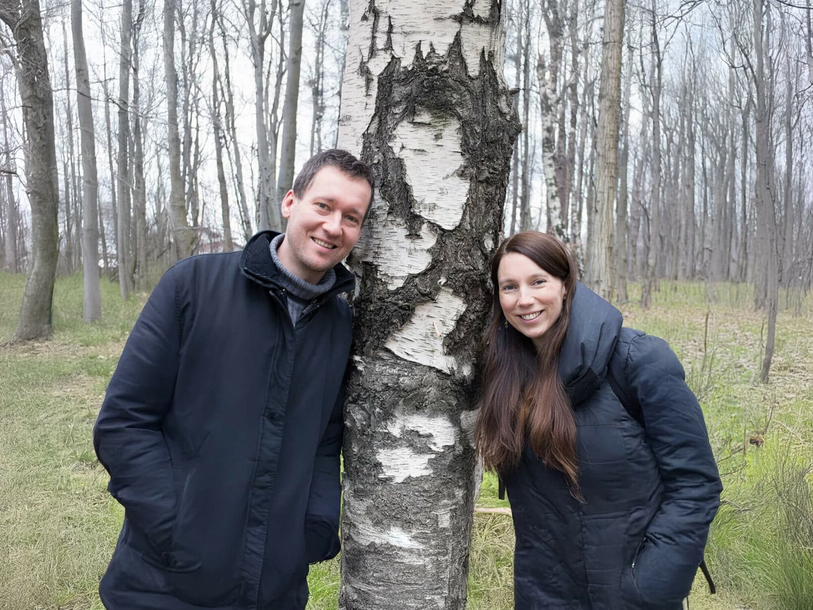 Paar in Winterjacken lehnt an Birkenbaum im Wald.